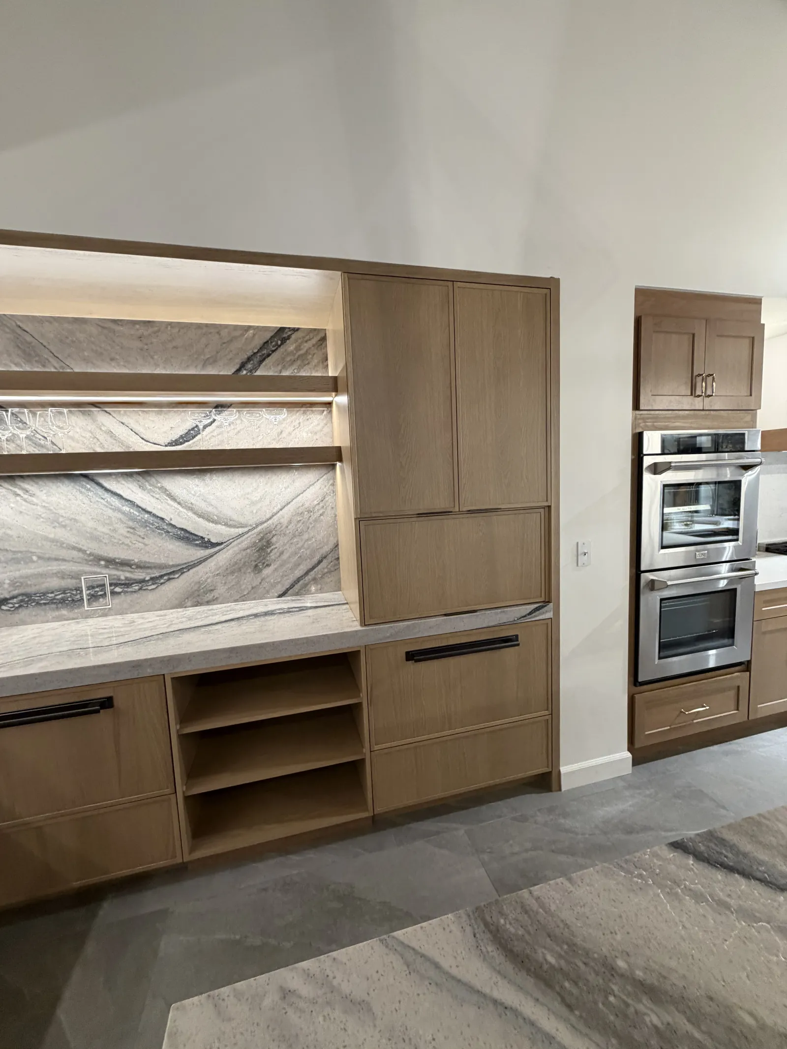 Eclipse Napa door style on the wine wall — open floating shelves with stem rails, marble back, and a flush-mount double oven in White Oak Yosemite Brown