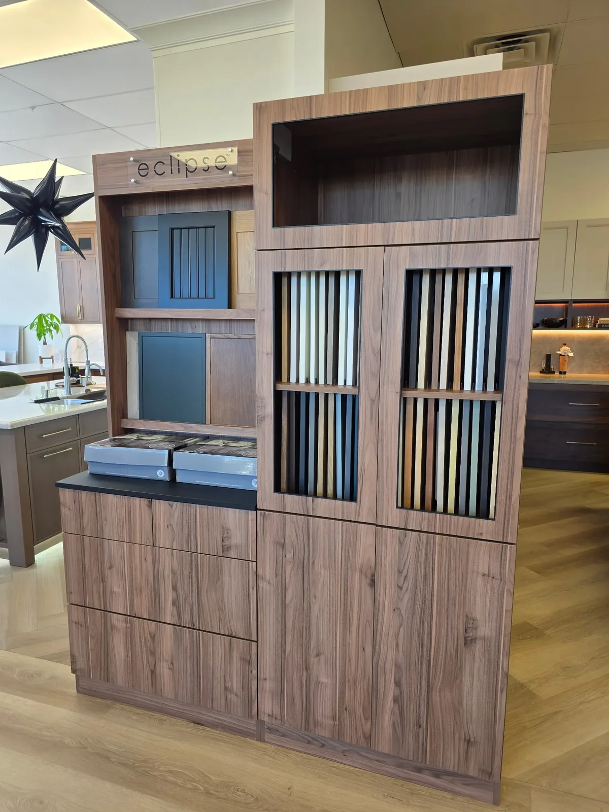 Eclipse Merchandiser display in a dealer showroom — walnut-finished hutch with the eclipse sign at top, glass-framed display cabinets holding vertical Eclipse door samples and finish chips, low credenza base with branded sample books on top
