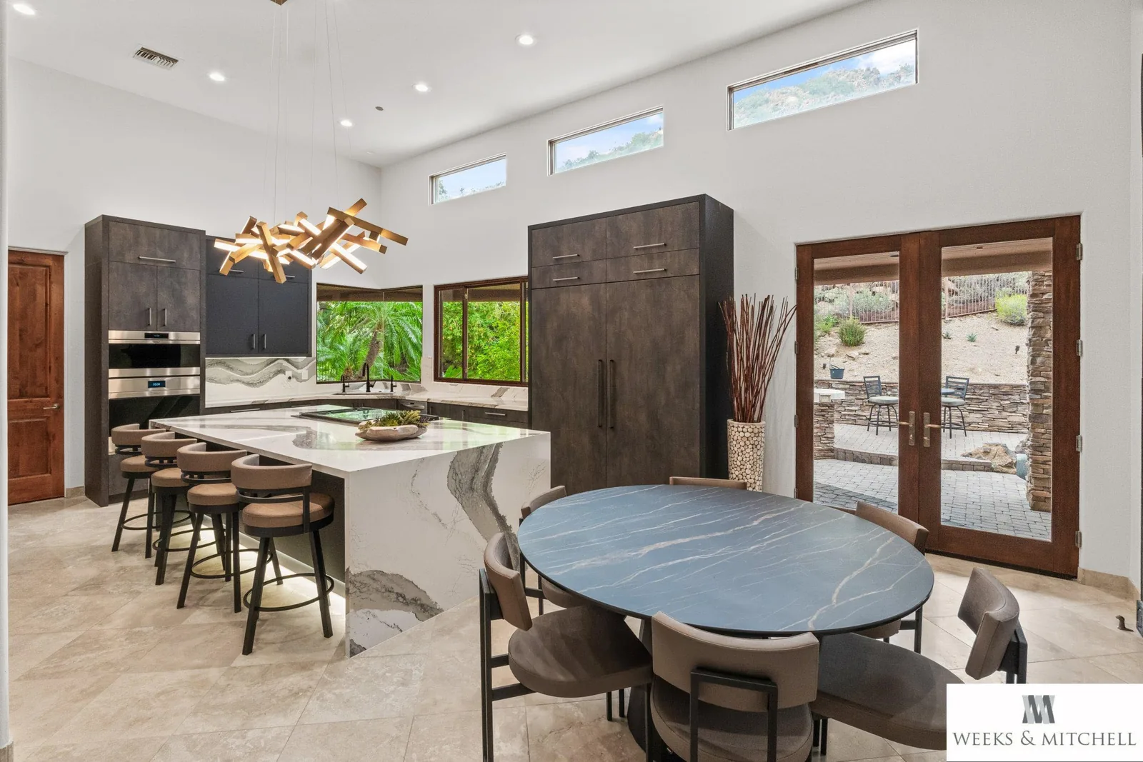 Dining area beside the Pronorm kitchen island with a Dark Steel pantry column and French doors opening to a paver patio and desert landscape