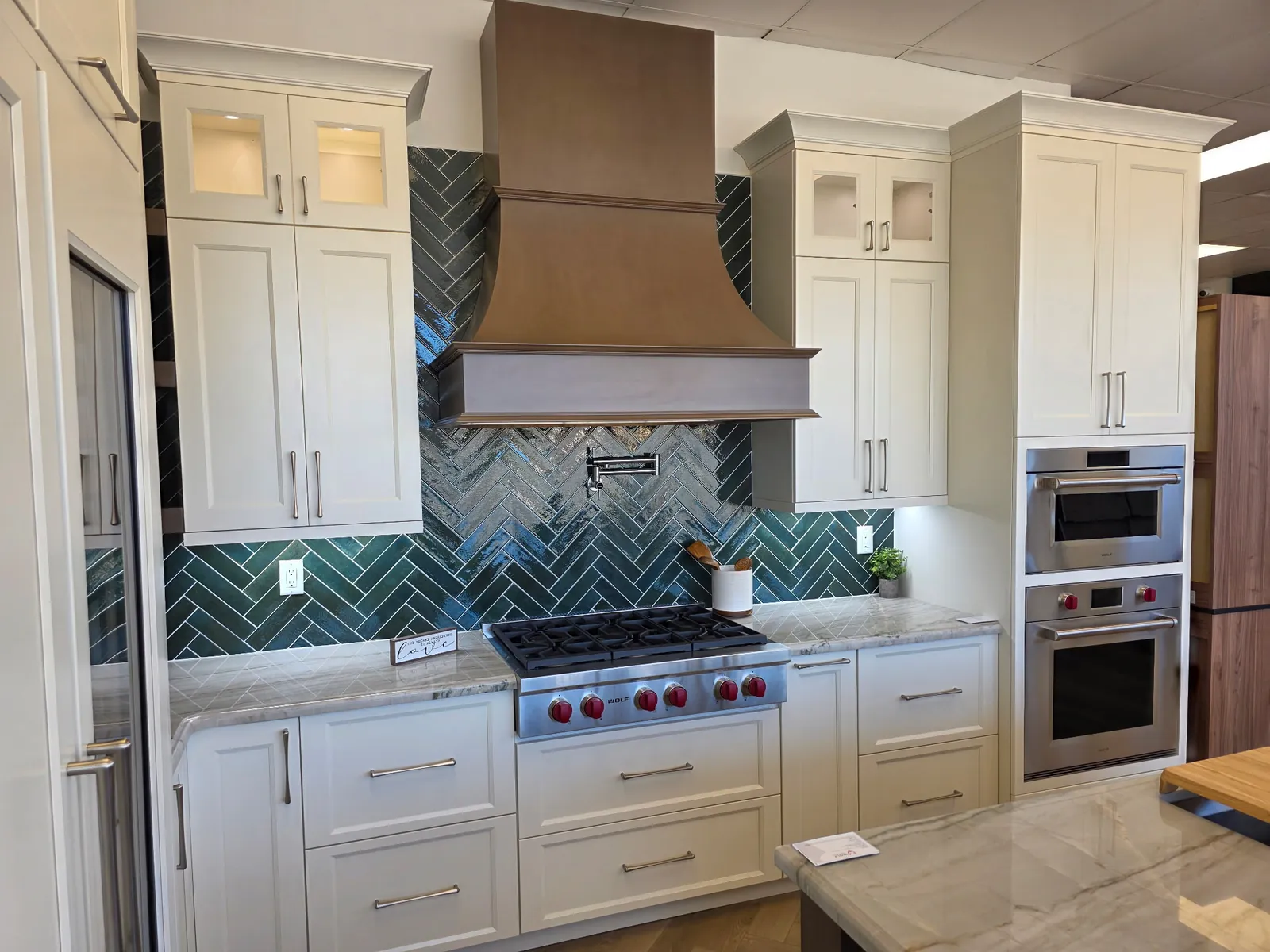 Wolf range and copper-bronze hood against the deep green herringbone backsplash, framed by Shiloh Statesville cabinetry in Shoji White