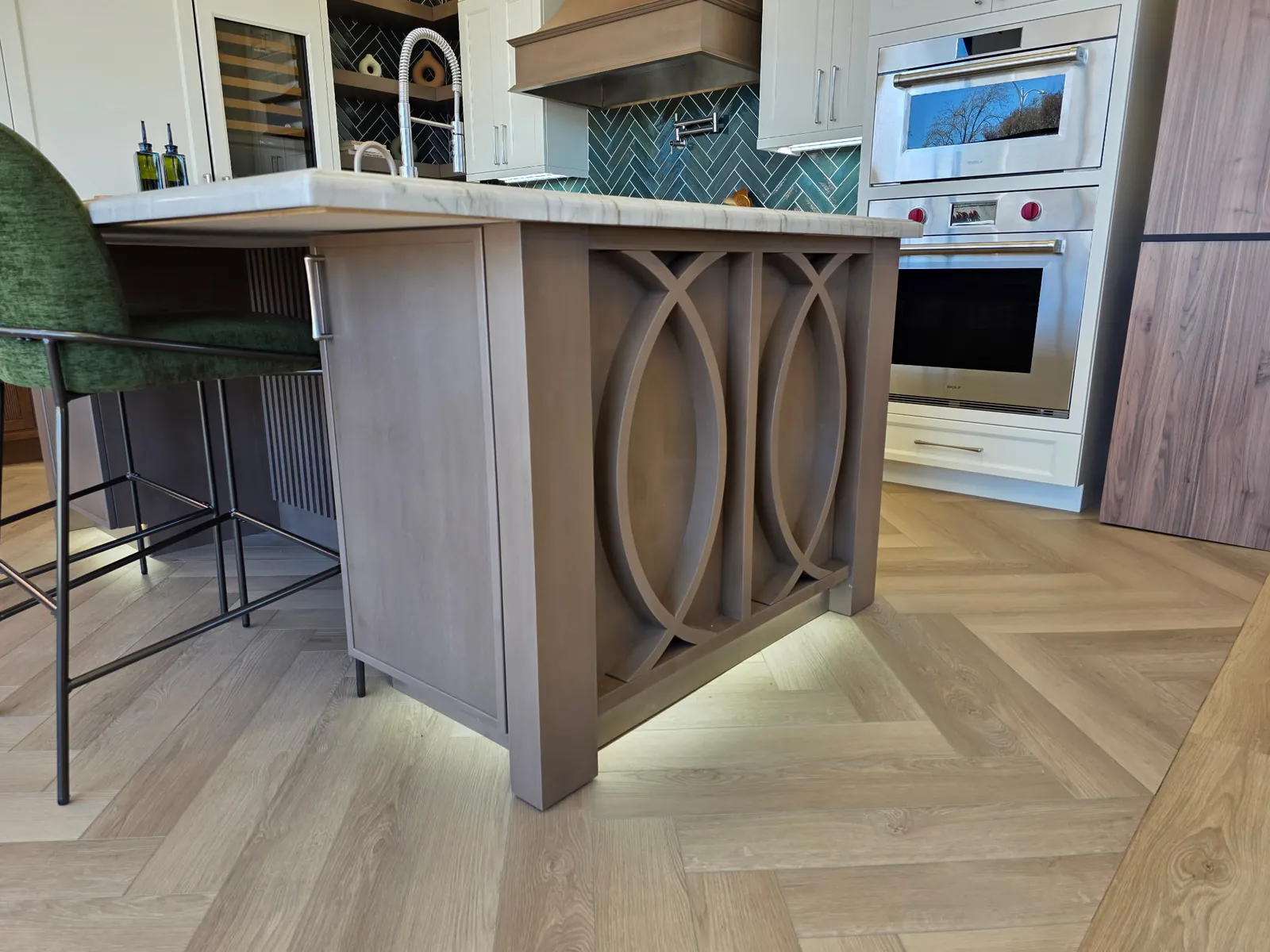 Greige (warm grey-brown) island with decorative oval panel detail on the end, Wolf wall oven and Sub-Zero column visible behind, herringbone wood floor