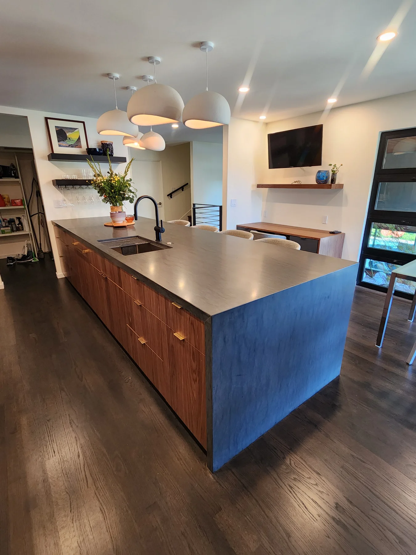 Distinctive Cabinetry Walnut Creek kitchen — island side detail with Eclipse Natural Walnut Metro fronts, brass flat-bar pulls, a deep-blue painted end-return panel, honed concrete-look counter, undermount prep sink, and a black articulating faucet
