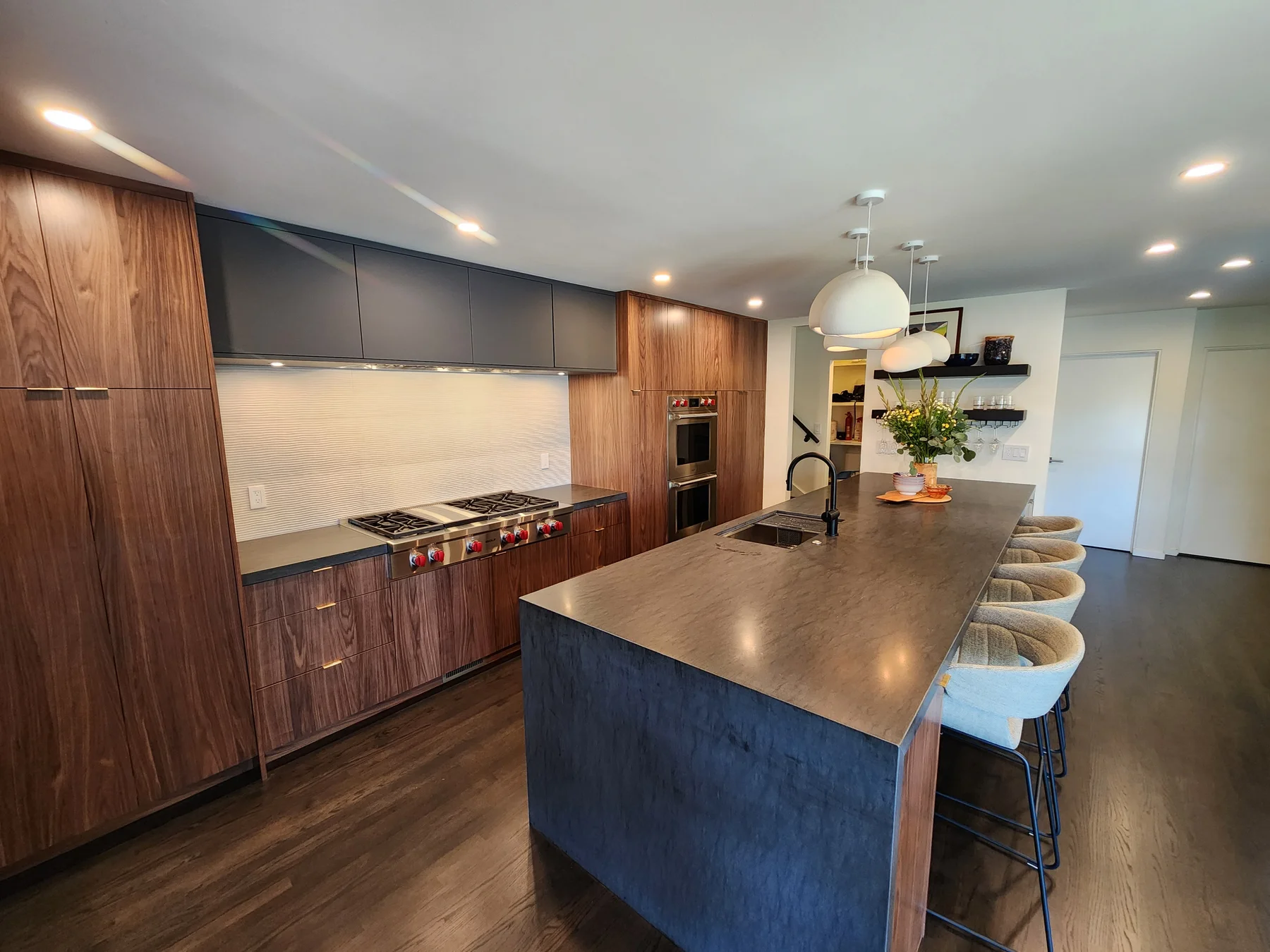 Distinctive Cabinetry Walnut Creek kitchen — Wolf rangetop wall with paneled walnut vent hood, matte Smoke Stack HPL flat-front uppers, textured white tile back, and adjacent walnut Wolf double wall ovens; the deep-blue island return visible across the room