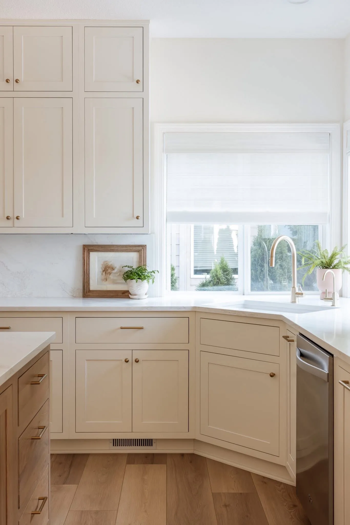 Stutler Cabinets Hanover kitchen — corner detail with brass-faucet sink under window, Soft White Hanover doors, marble backsplash, framed art