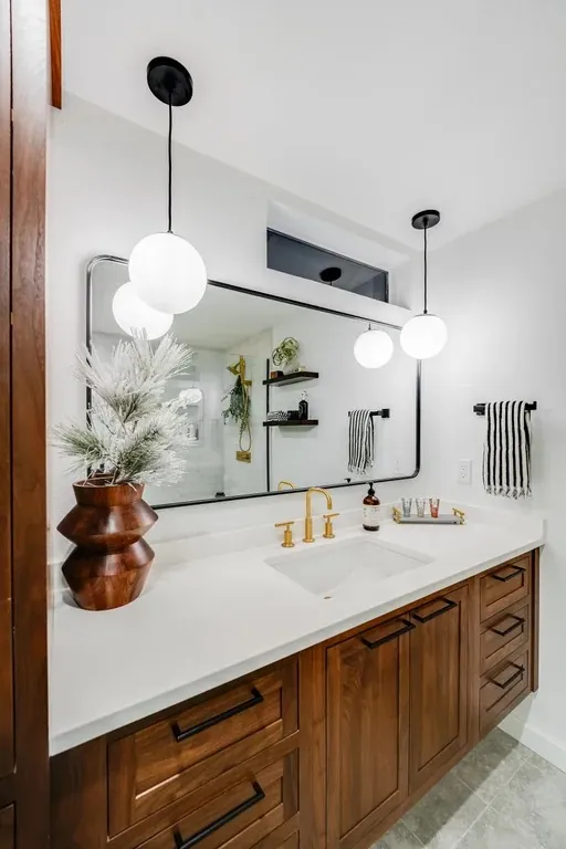 Stutler Cabinets master bath — straight-on view of the vanity with brass faucet, pampas in a turned wood vase, frameless shower visible in the mirror